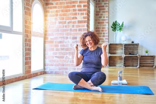 Middle age beautiful sportswoman wearing sportswear sitting on mat practicing yoga at home very happy and excited doing winner gesture with arms raised, smiling and screaming for success. Celebration