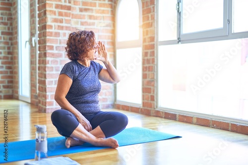 Middle age beautiful sportswoman wearing sportswear sitting on mat practicing yoga at home shouting and screaming loud to side with hand on mouth. Communication concept.