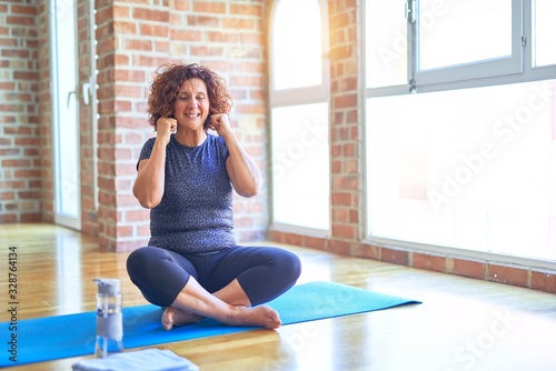 Middle age beautiful sportswoman wearing sportswear sitting on mat practicing yoga at home covering ears with fingers with annoyed expression for the noise of loud music. Deaf concept.