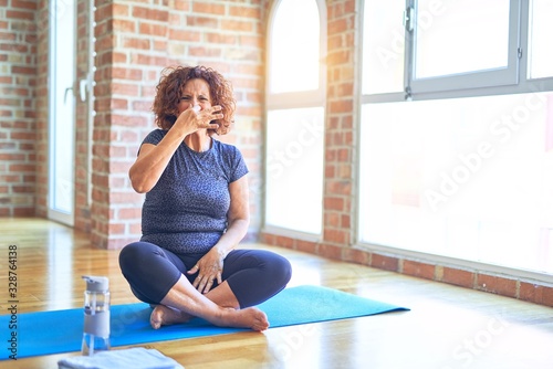 Middle age beautiful sportswoman wearing sportswear sitting on mat practicing yoga at home smelling something stinky and disgusting, intolerable smell, holding breath with fingers on nose. Bad smells