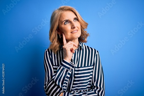 Middle age beautiful blonde woman wearing casual striped shirt standing over blue background with hand on chin thinking about question, pensive expression. Smiling with thoughtful face. Doubt concept.