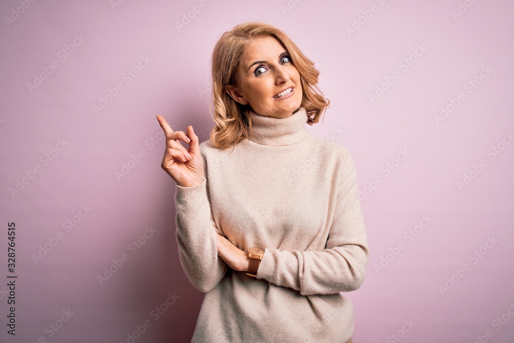 Middle age beautiful blonde woman wearing casual turtleneck sweater over pink background with a big smile on face, pointing with hand and finger to the side looking at the camera. Middle age beautiful blonde woman wearing casual turtleneck sweater over pink background with a big smile on face, pointing with hand and finger to the side looking at the camera.
