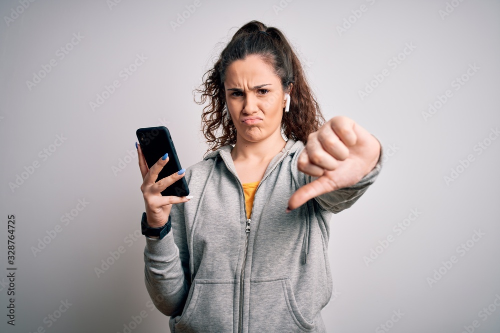 Naklejka premium Beautiful sportswoman with curly hair listening to music using smartphone and earphones with angry face, negative sign showing dislike with thumbs down, rejection concept