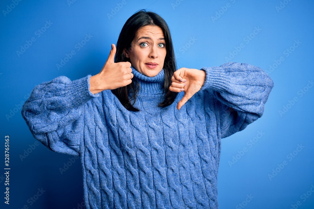 Young brunette woman with blue eyes wearing casual turtleneck sweater Doing thumbs up and down, disagreement and agreement expression. Crazy conflict