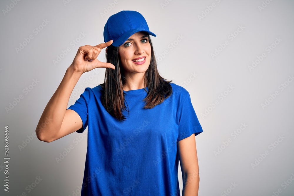 Young delivery woman with blue eyes wearing cap standing over blue background smiling and confident gesturing with hand doing small size sign with fingers looking and the camera. Measure concept. Young delivery woman with blue eyes wearing cap standing over blue background smiling and confident gesturing with hand doing small size sign with fingers looking and the camera. Measure concept.