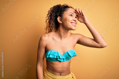 Young beautiful american woman on vacation wearing bikini over isolated yellow background very happy and smiling looking far away with hand over head. Searching concept.