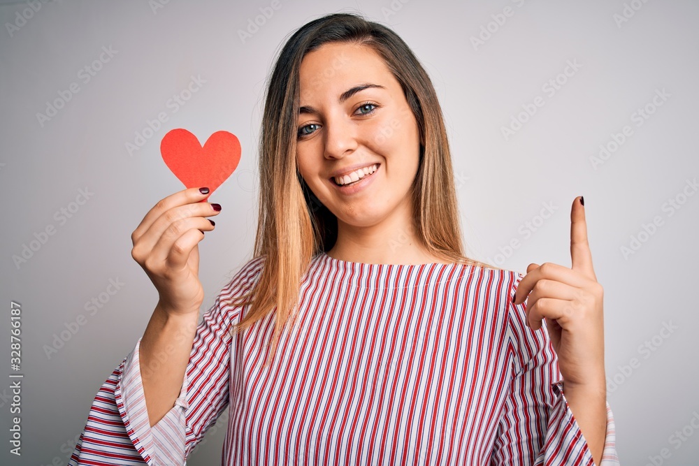 Young beautiful blonde romantic woman holding red paper heart over white background surprised with an idea or question pointing finger with happy face, number one Young beautiful blonde romantic woman holding red paper heart over white background surprised with an idea or question pointing finger with happy face, number one