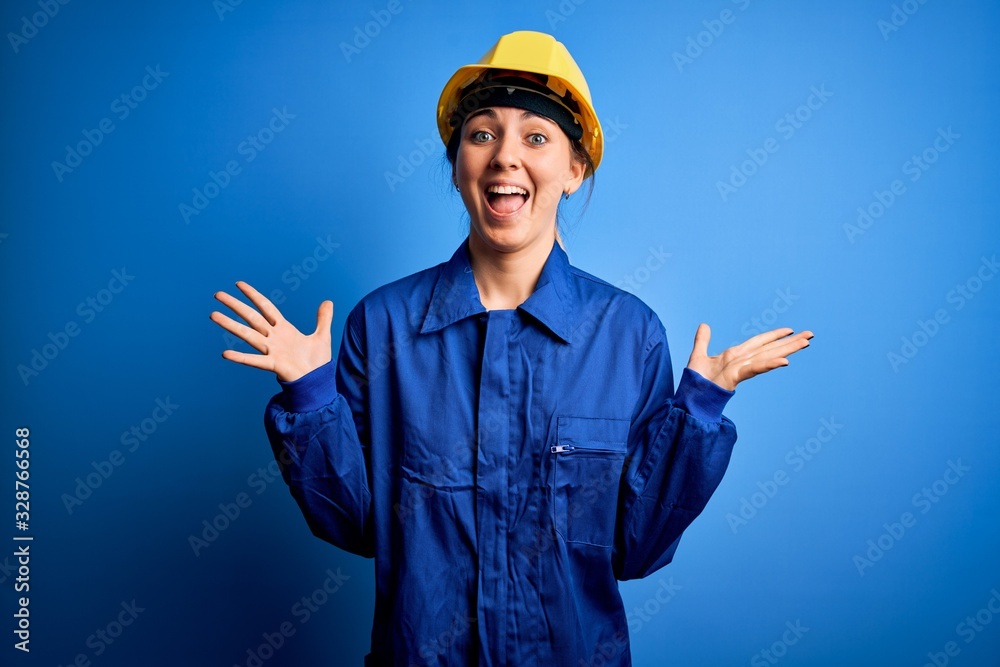 Young beautiful worker woman with blue eyes wearing security helmet and uniform celebrating crazy and amazed for success with arms raised and open eyes screaming excited. Winner concept