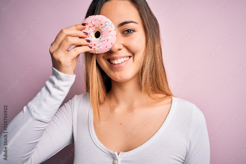 Young beautiful blonde woman with blue eyes holding pink doughnut over isolated background with a happy face standing and smiling with a confident smile showing teeth Young beautiful blonde woman with blue eyes holding pink doughnut over isolated background with a happy face standing and smiling with a confident smile showing teeth