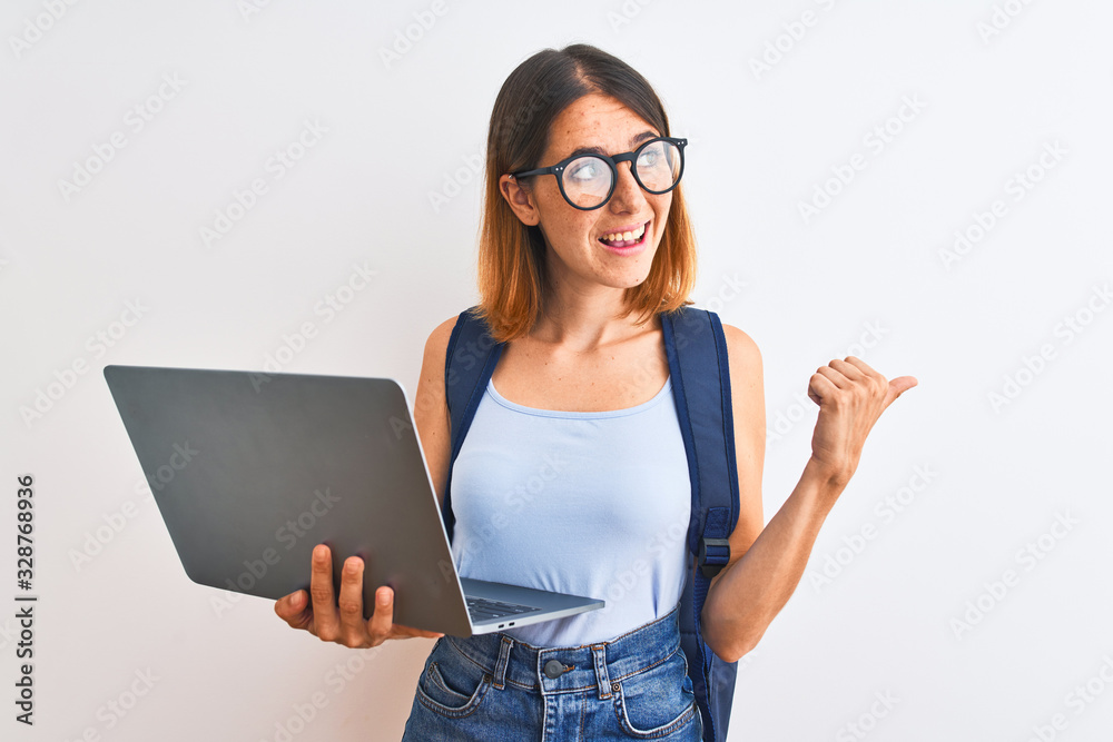 Beautiful redhead student woman wearing a backpack and using laptop pointing and showing with thumb up to the side with happy face smiling