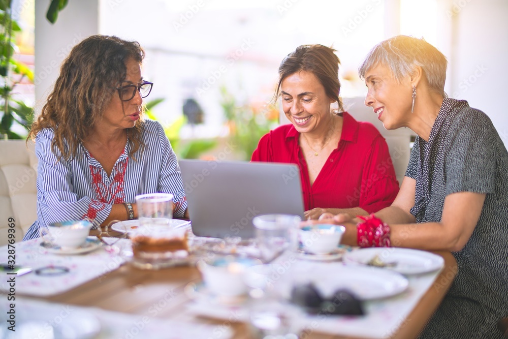 Meeting of middle age women having lunch and drinking coffee. Mature friends smiling happy using laptop at home on a sunny day