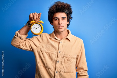 Young handsome man holding alarm clock standing over isolated blue background with a confident expression on smart face thinking serious
