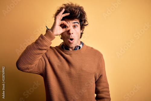 Young handsome man wearing casual shirt and sweater over isolated yellow background doing ok gesture shocked with surprised face, eye looking through fingers. Unbelieving expression.