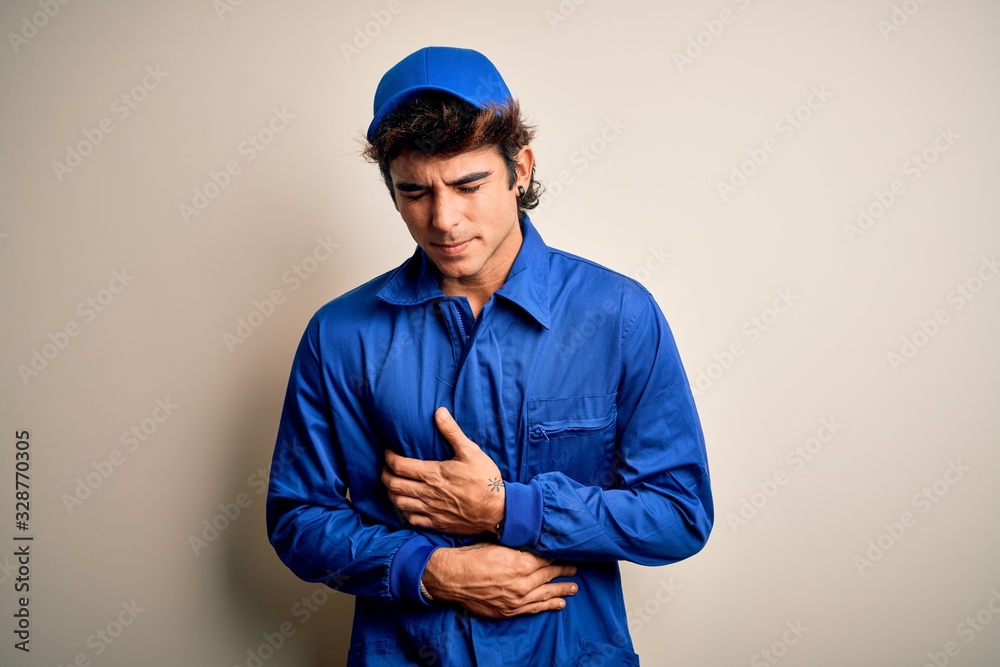 Young mechanic man wearing blue cap and uniform standing over isolated white background with hand on stomach because indigestion, painful illness feeling unwell. Ache concept.