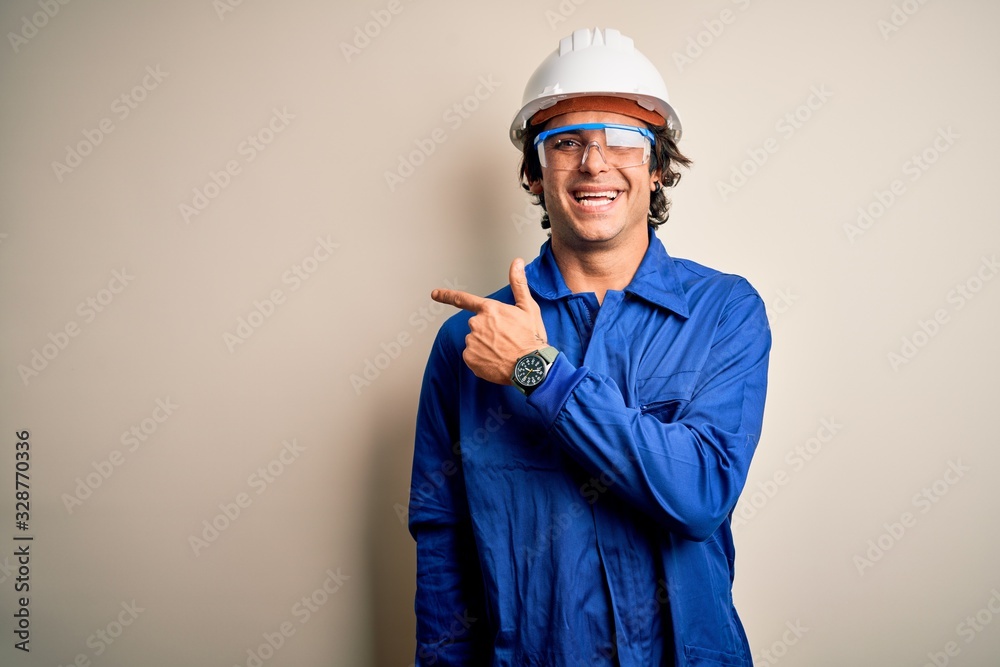 Young constructor man wearing uniform and security helmet over isolated white background cheerful with a smile of face pointing with hand and finger up to the side with happy and natural expression