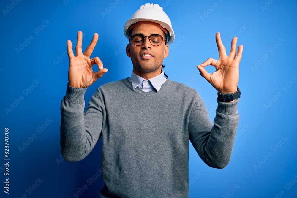 Young handsome african american engineer man with dreadlocks wearing safety helmet relax and smiling with eyes closed doing meditation gesture with fingers. Yoga concept. Young handsome african american engineer man with dreadlocks wearing safety helmet relax and smiling with eyes closed doing meditation gesture with fingers. Yoga concept.