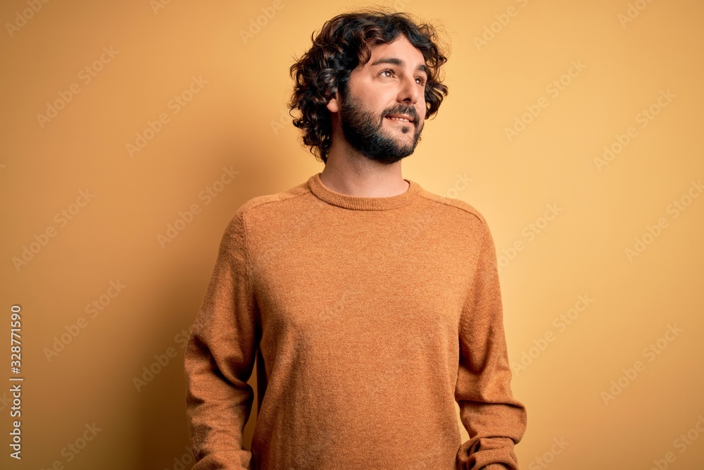 Obraz premium Young handsome man with beard wearing casual sweater standing over yellow background looking away to side with smile on face, natural expression. Laughing confident.