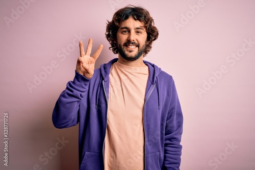 Young handsome sporty man with beard wearing casual sweatshirt over pink background showing and pointing up with fingers number three while smiling confident and happy.