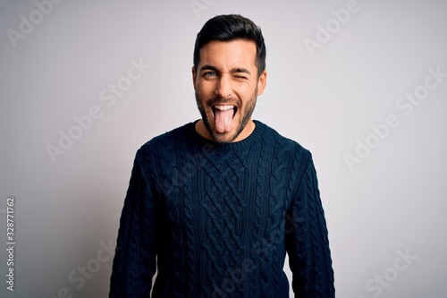 Young handsome man with beard wearing casual sweater standing over white background sticking tongue out happy with funny expression. Emotion concept. photo
