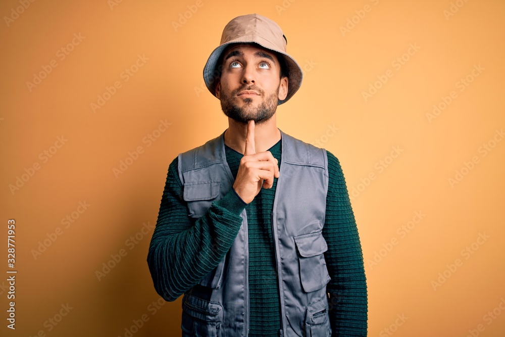 Handsome tourist man with beard on vacation wearing explorer hat over yellow background Thinking concentrated about doubt with finger on chin and looking up wondering
