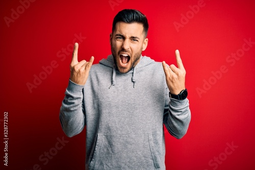 Tableau sur toile Young handsome sportsman wearing sweatshirt standing over isolated red background shouting with crazy expression doing rock symbol with hands up