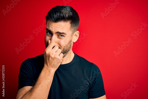 Young handsome man wearing casual black t-shirt standing over isolated red background smelling something stinky and disgusting, intolerable smell, holding breath with fingers on nose. Bad smell