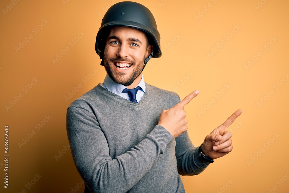 Young handsome businessman wearing military helmet over isolated yellow background smiling and looking at the camera pointing with two hands and fingers to the side. Young handsome businessman wearing military helmet over isolated yellow background smiling and looking at the camera pointing with two hands and fingers to the side.