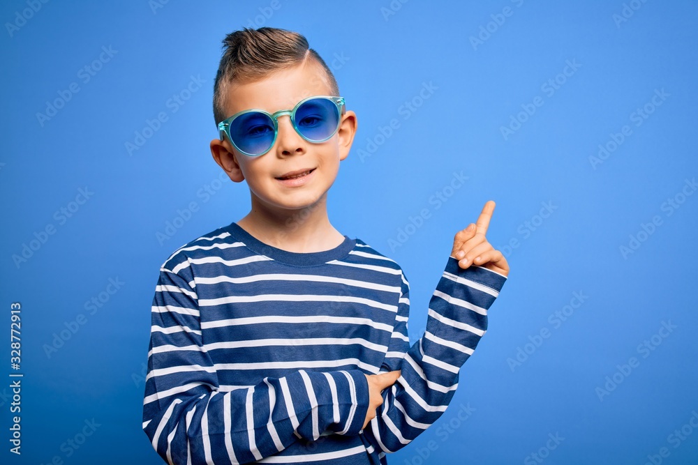 Young little caucasian kid with blue eyes standing wearing sunglasses over blue background with a big smile on face, pointing with hand and finger to the side looking at the camera. Young little caucasian kid with blue eyes standing wearing sunglasses over blue background with a big smile on face, pointing with hand and finger to the side looking at the camera.
