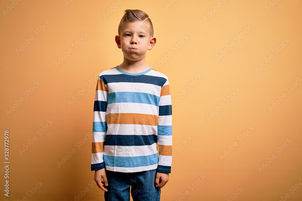 Young little caucasian kid with blue eyes wearing colorful striped shirt over yellow background puffing cheeks with funny face. Mouth inflated with air, crazy expression. Young little caucasian kid with blue eyes wearing colorful striped shirt over yellow background puffing cheeks with funny face. Mouth inflated with air, crazy expression.