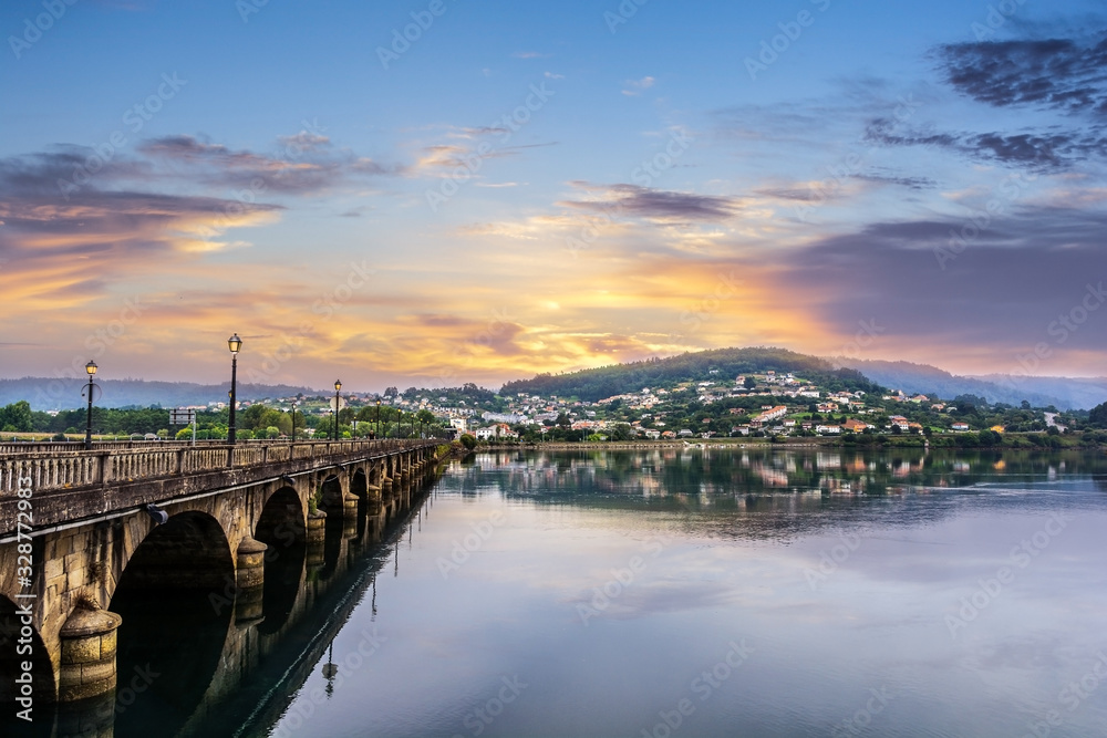 Fototapeta premium Sunset in Pontedeume, Galicia, Spain Ancient Roman stone bridge and reflections of the hill and village in the calm water. Golden clouds and colors of the sunset