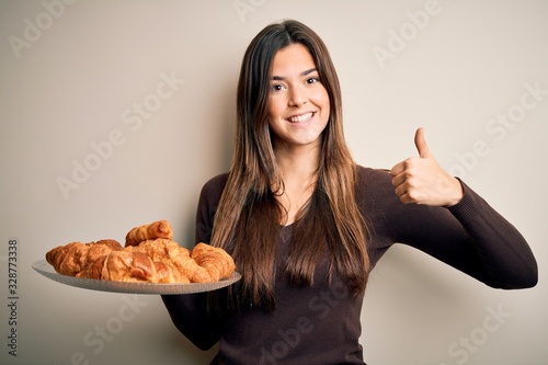 Young beautiful girl holding plate with sweet croissants for breakfast over white background happy with big smile doing ok sign  thumb up with fingers  excellent sign
