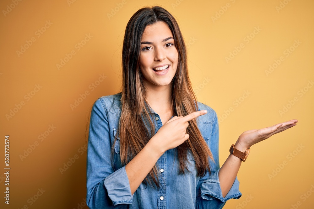 Fototapeta premium Young beautiful girl wearing casual denim shirt standing over isolated yellow background amazed and smiling to the camera while presenting with hand and pointing with finger.