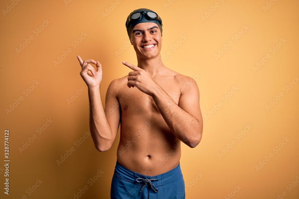 Young handsome man shirtless wearing swimsuit and swim cap over isolated yellow background smiling and looking at the camera pointing with two hands and fingers to the side.