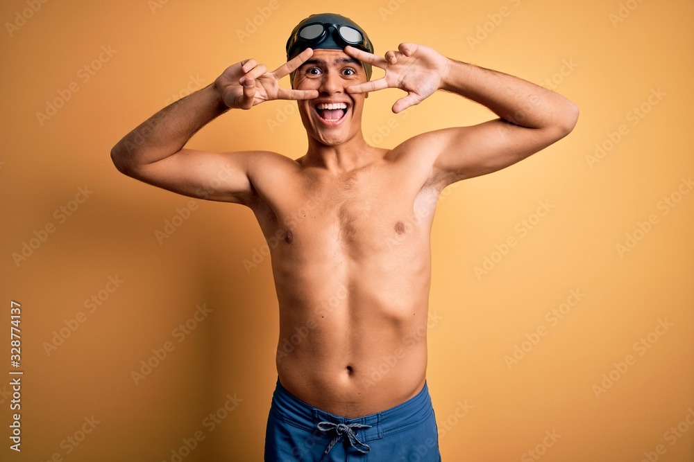 Young handsome man shirtless wearing swimsuit and swim cap over isolated yellow background Doing peace symbol with fingers over face, smiling cheerful showing victory