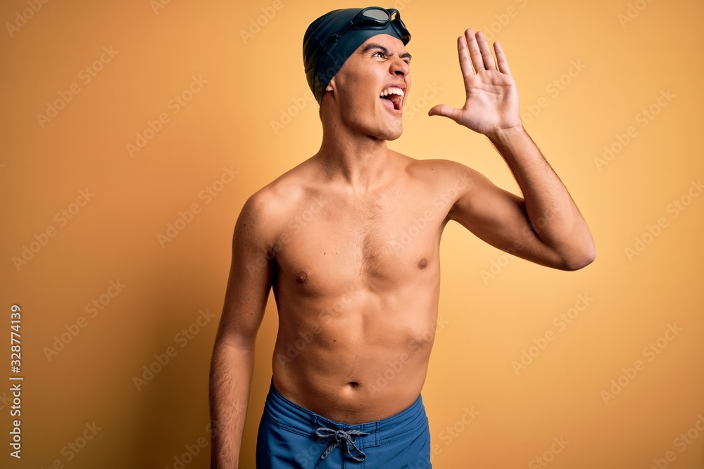 Young handsome man shirtless wearing swimsuit and swim cap over isolated yellow background shouting and screaming loud to side with hand on mouth. Communication concept.