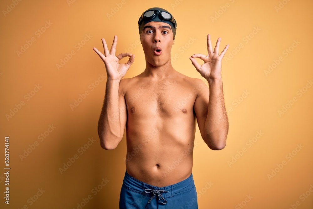 Young handsome man shirtless wearing swimsuit and swim cap over isolated yellow background looking surprised and shocked doing ok approval symbol with fingers. Crazy expression