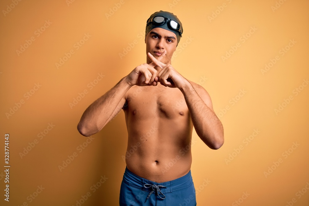 Young handsome man shirtless wearing swimsuit and swim cap over isolated yellow background Rejection expression crossing fingers doing negative sign