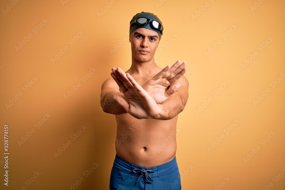 Young handsome man shirtless wearing swimsuit and swim cap over isolated yellow background Rejection expression crossing arms and palms doing negative sign, angry face