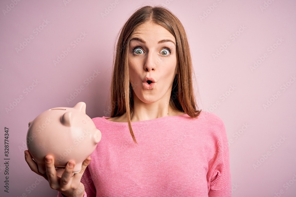 Young beautiful redhead woman holding piggy bank over isolated pink background scared in shock with a surprise face, afraid and excited with fear expression