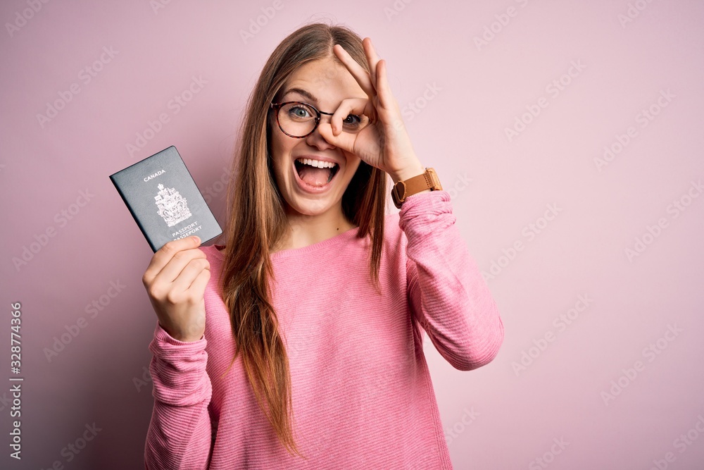 Beautiful redhead tourist woman holding canadian canada passport over pink bakcground with happy face smiling doing ok sign with hand on eye looking through fingers