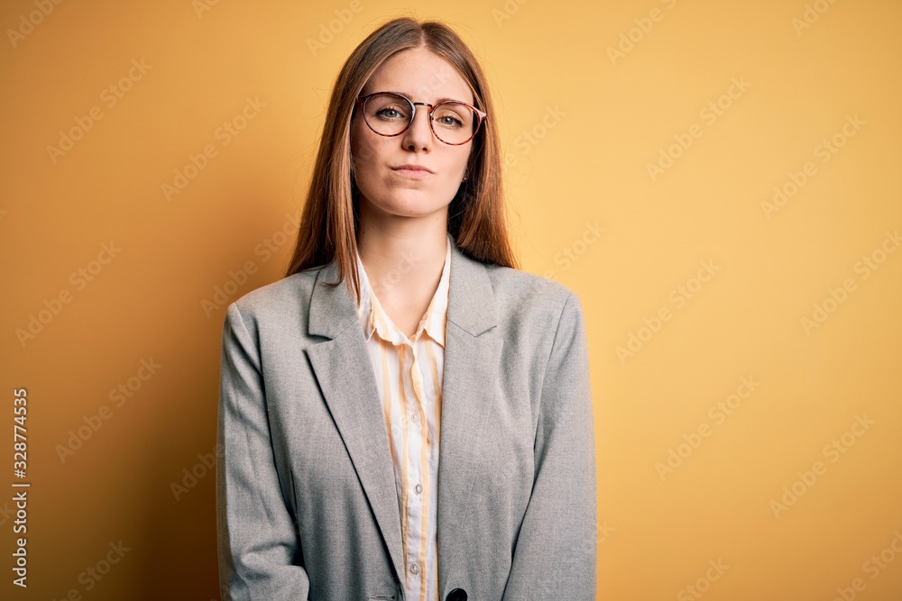 Young beautiful redhead woman wearing jacket and glasses over isolated yellow background Relaxed with serious expression on face. Simple and natural looking at the camera.
