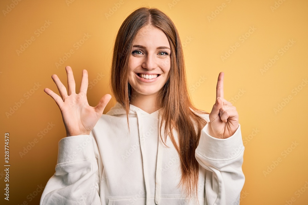 Young beautiful redhead sporty woman wearing sweatshirt over isolated yellow background showing and pointing up with fingers number six while smiling confident and happy. Young beautiful redhead sporty woman wearing sweatshirt over isolated yellow background showing and pointing up with fingers number six while smiling confident and happy.