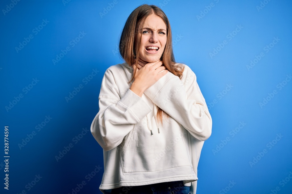 Young beautiful redhead sporty woman wearing sweatshirt over isolated blue background shouting and suffocate because painful strangle. Health problem. Asphyxiate and suicide concept.