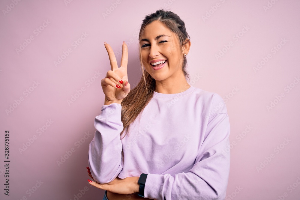 Young beautiful sport woman wearing sweatshirt over pink isolated background smiling with happy face winking at the camera doing victory sign. Number two. Young beautiful sport woman wearing sweatshirt over pink isolated background smiling with happy face winking at the camera doing victory sign. Number two.