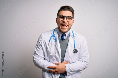 Young doctor man with blue eyes wearing medical coat and stethoscope over isolated background smiling and laughing hard out loud because funny crazy joke with hands on body.