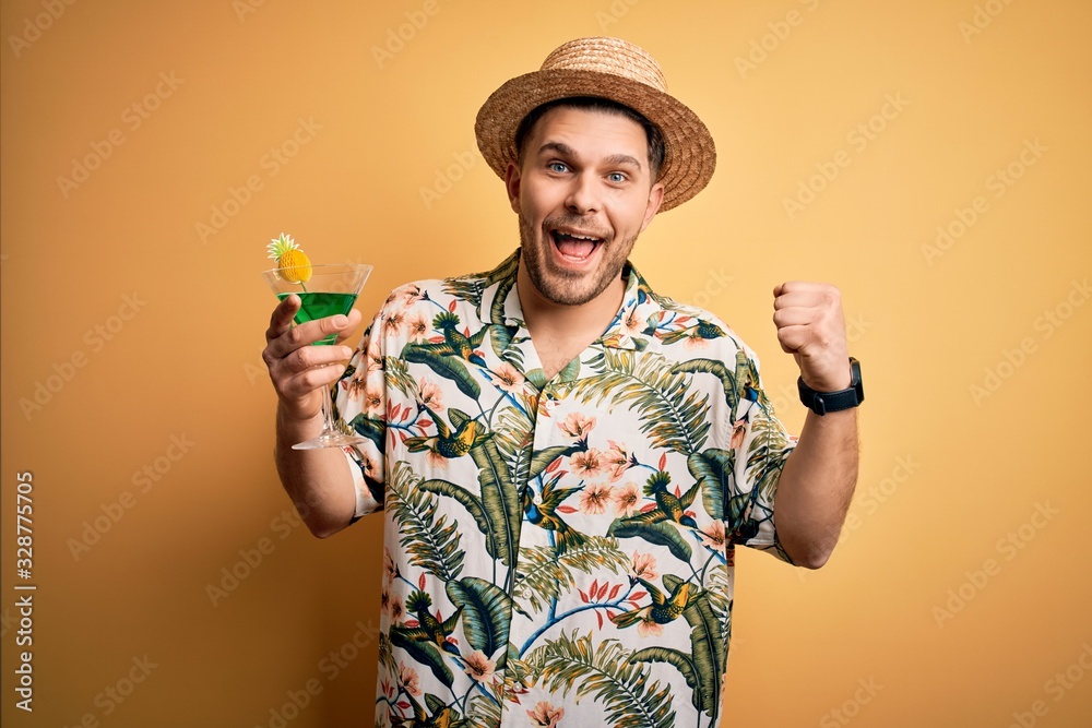 Young man on vacation wearing summer hat drinking a party cocktail over yellow background screaming proud and celebrating victory and success very excited, cheering emotion Young man on vacation wearing summer hat drinking a party cocktail over yellow background screaming proud and celebrating victory and success very excited, cheering emotion