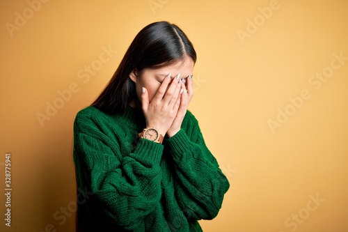 Young beautiful asian woman wearing green winter sweater over yellow isolated background with sad expression covering face with hands while crying. Depression concept. © Krakenimages.com