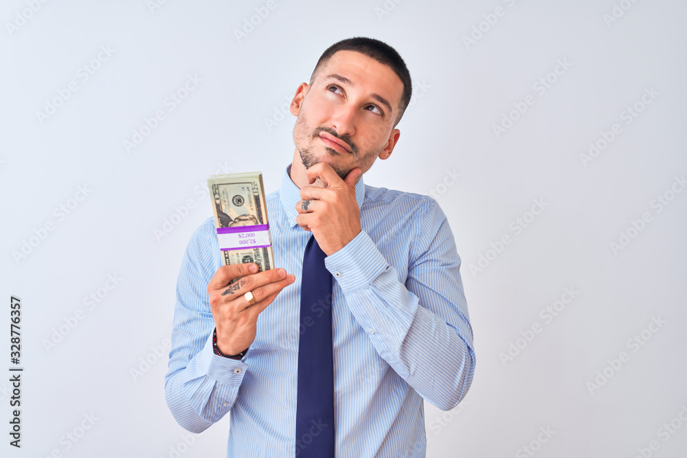 Young handsome business man holding a bunch of dollars bank notes over isolated background serious face thinking about question, very confused idea Young handsome business man holding a bunch of dollars bank notes over isolated background serious face thinking about question, very confused idea