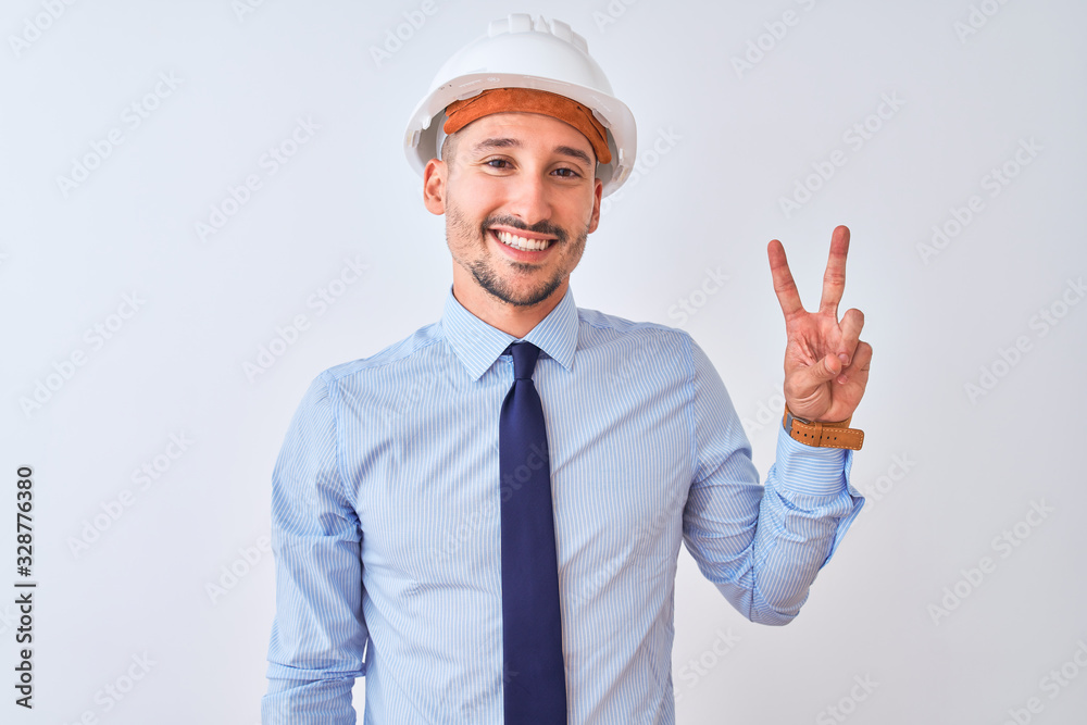 Young business man wearing contractor safety helmet over isolated background smiling with happy face winking at the camera doing victory sign. Number two. Young business man wearing contractor safety helmet over isolated background smiling with happy face winking at the camera doing victory sign. Number two.