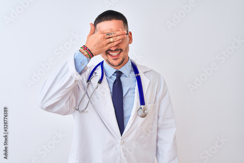 Young doctor man wearing stethoscope over isolated background smiling and laughing with hand on face covering eyes for surprise. Blind concept.